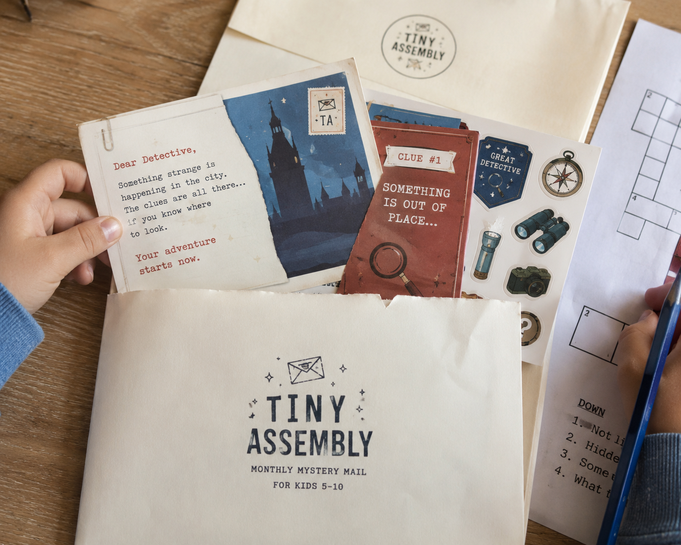 Tiny Assembly monthly mystery mail — envelope, story letter, clue cards, stickers and puzzle spread on a wooden table